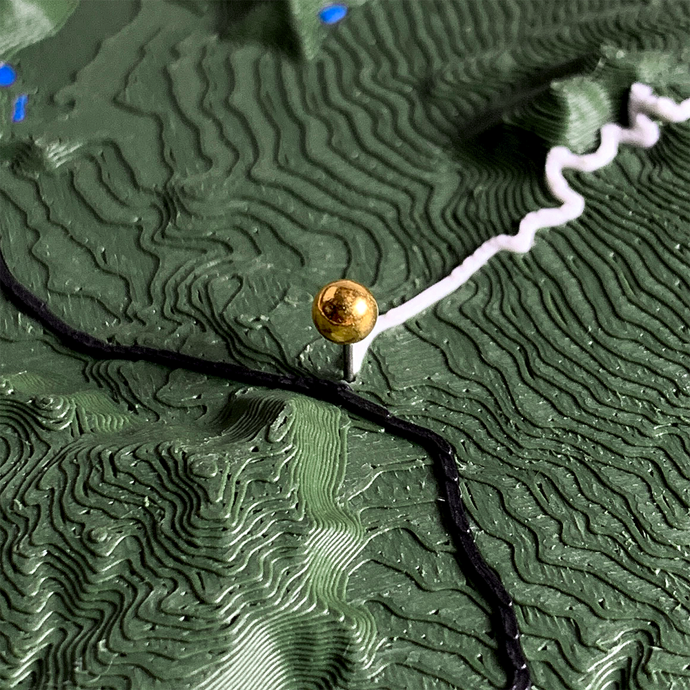 Close-up of a 3D printed terrain relief in dark green, showing a polished gold
metal location marker pin set into a hillside where a black route line and a
white route line meet. Fine print layers form contour ridges across the slope.