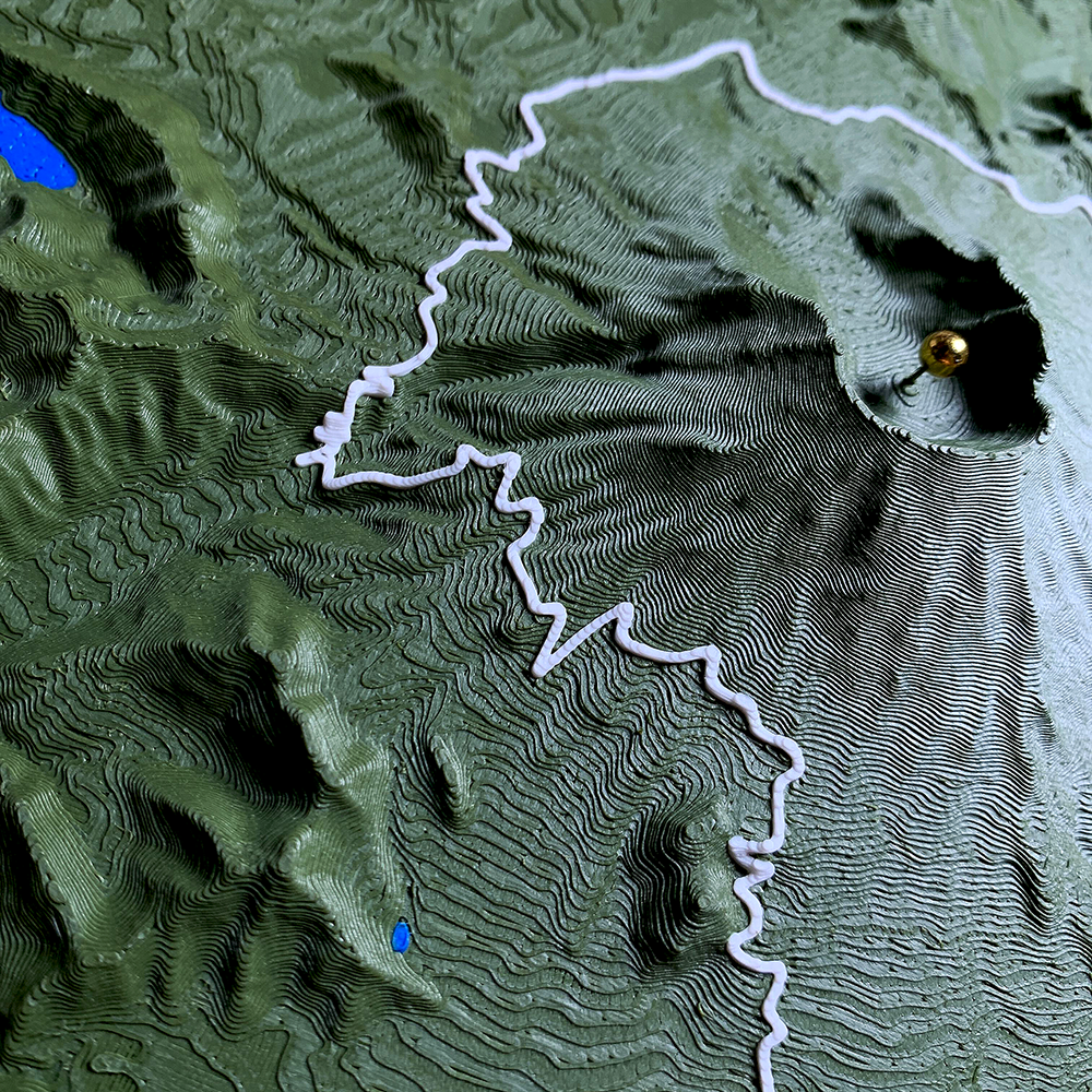 Wide view of a 3D printed topographic relief in dark green, with a white route
line switchbacking up a steep valley toward a gold marker pin on the far ridge.
Small blue lakes sit in the basins below and print layers trace every contour.
