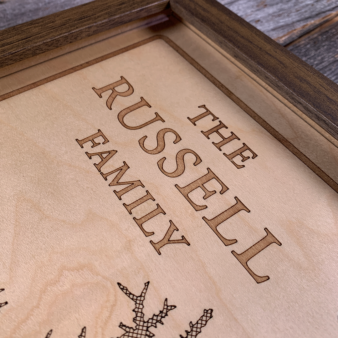Close-up of laser-engraved family name on framed wood map artwork. Precision engraving on maple highlights personalized text, showcasing craftsmanship and custom detail for meaningful lake house décor.