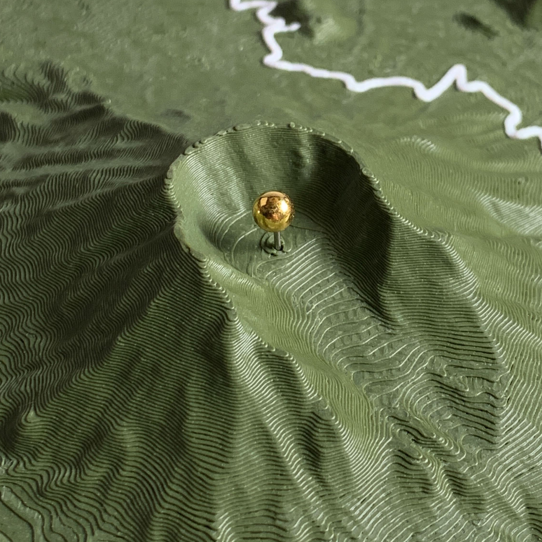 Gold metal marker pin set inside the horseshoe crater of a 3D printed Mount
Saint Helens terrain relief in dark green, with print layers fanning down the
steep crater walls and a white route line crossing the ridge above.