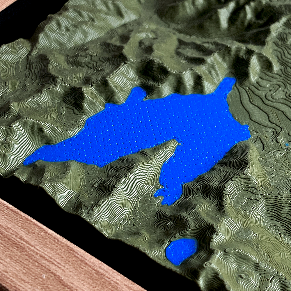 Macro detail of a 3D printed topographic relief in dark green, with individual
print layers tracing the contours of steep valley walls around a bright blue
lake. The edge of the wood frame is visible at the lower left.