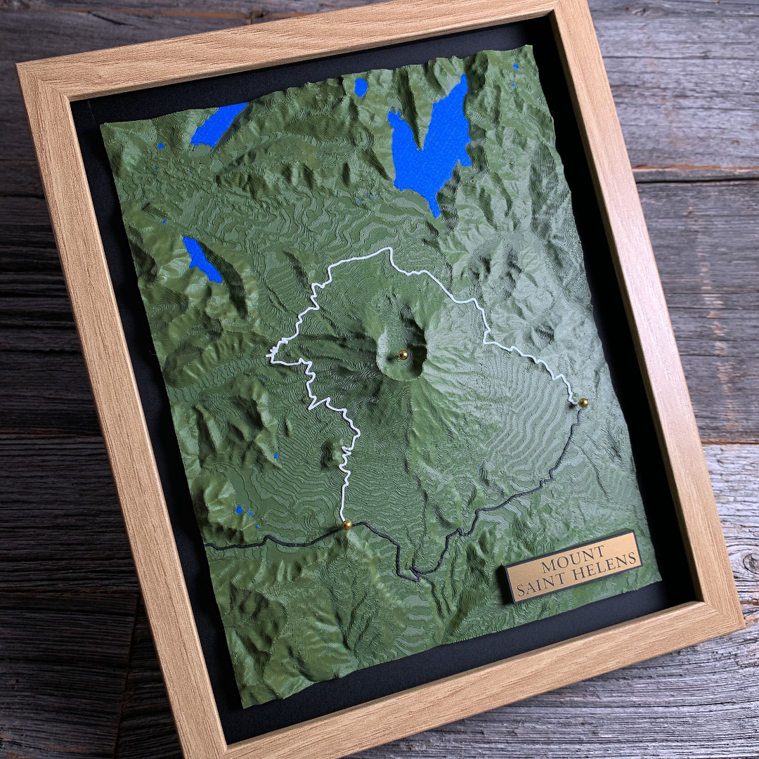 Complete framed 3D Terrain Art piece of Mount Saint Helens, Washington. Dark
green printed relief with the volcano's breached crater at centre, blue lakes
to the north, a white loop trail and a black road traced across the terrain,
three gold marker pins, and a gold engraved title block reading "Mount Saint
Helens." Shown in a light oak frame on weathered grey wood.