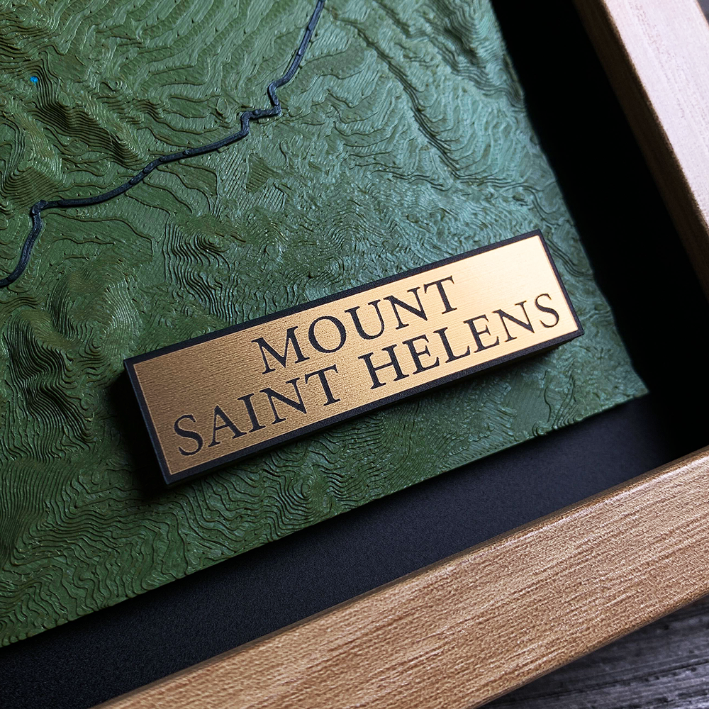 Gold engraved title block reading "Mount Saint Helens" mounted on a raised
platform in a dark green 3D printed terrain relief, with a black route line
crossing the slope above and a light wood frame edge in the foreground.
